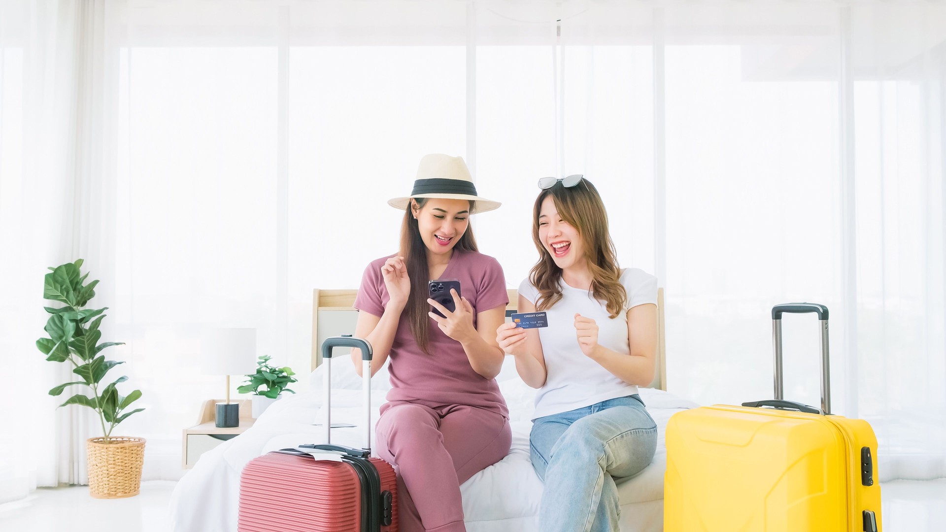 Two happy Asian women booked a hotel room on a smart phone and pay with a credit card while sitting together on the bed with their luggage. Planning on holiday vacation trip. Image with copy space.